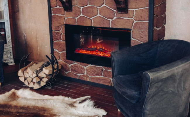 View over fireplace with burning logs, natural fur skin on the floor next to holder with logs in cozy room.